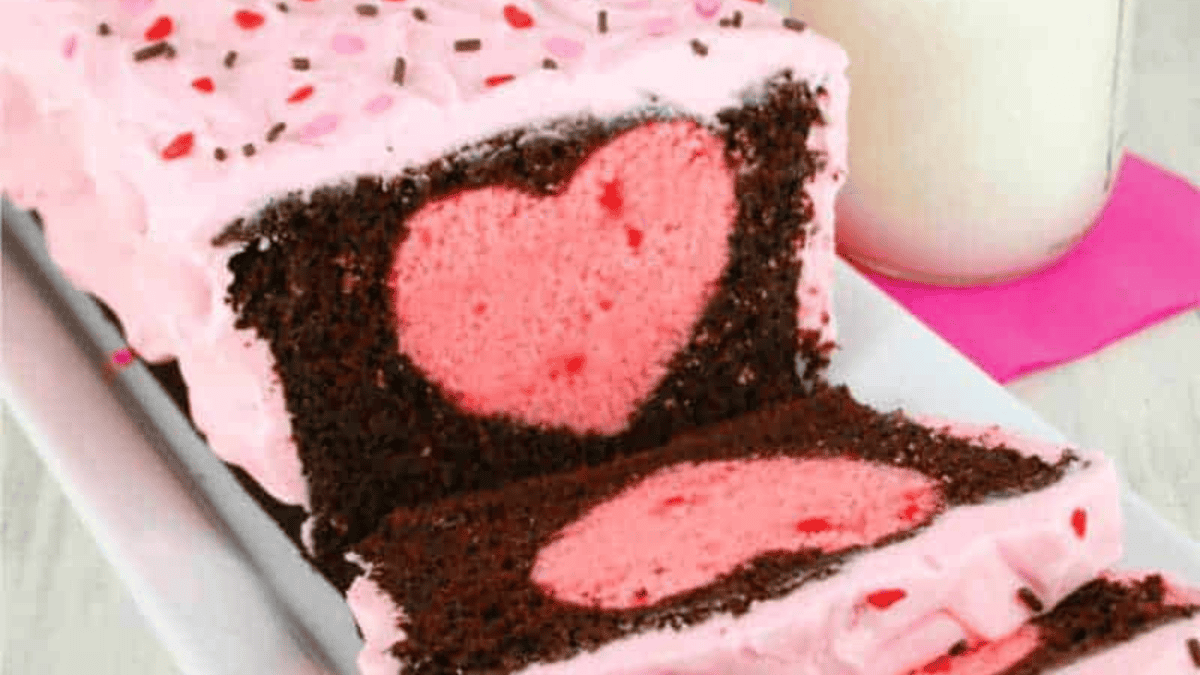 A chocolate loaf cake with pink frosting and sprinkles, sliced to reveal a pink heart shape in the center of each slice. A glass of milk sits in the background.