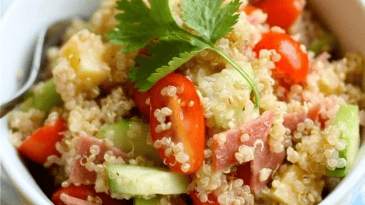 A bowl of quinoa salad with chopped cucumber, cherry tomatoes, ham pieces, and garnished with a sprig of cilantro.