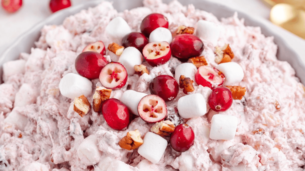 A close-up of a creamy salad topped with whole cranberries, mini marshmallows, and chopped pecans. The salad appears pink and fluffy, served in a white dish.