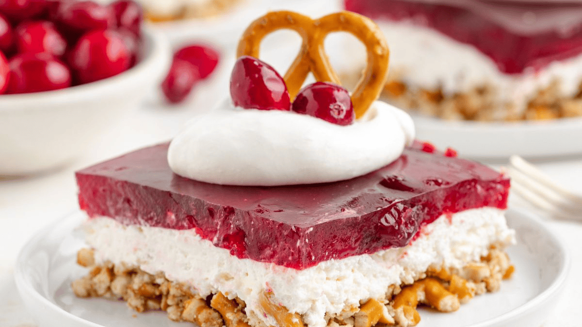 A slice of layered dessert with a pretzel crust, white creamy middle, red gelatin top, topped with whipped cream, two cherries, and a small pretzel. A bowl of cherries is visible in the background.