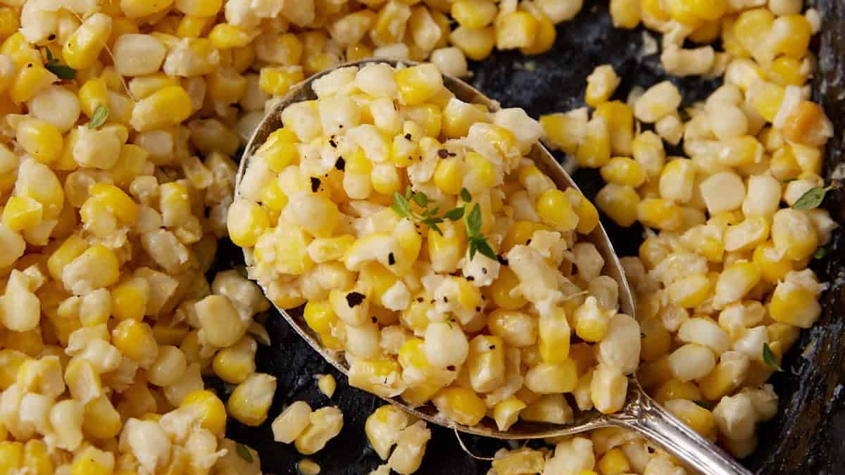 A close-up image shows a skillet filled with creamy corn. A silver spoon holds a portion of the creamed corn, revealing kernels cooked in a luscious sauce and garnished with fresh herbs. The corn looks well-seasoned and ready to serve.