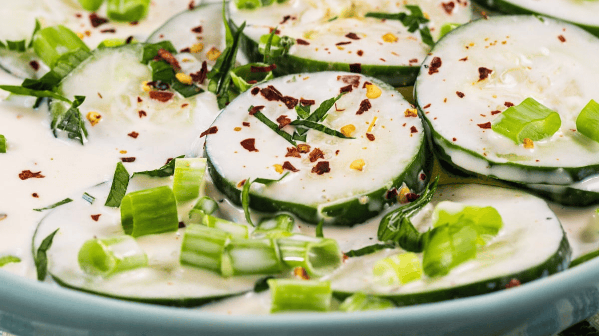Close-up of cucumber slices topped with creamy dressing, chopped green onions, fresh herbs, and red pepper flakes on a light blue plate.
