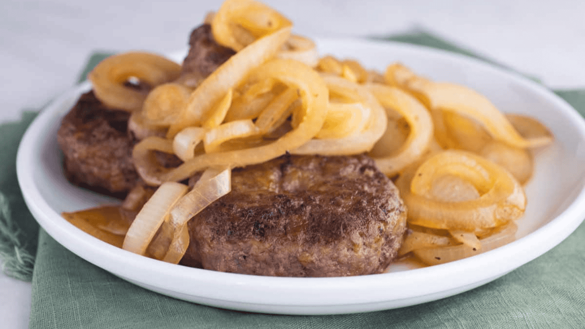A white plate holds browned hamburger steaks topped with sautéed sliced onions, placed on a green cloth napkin.
