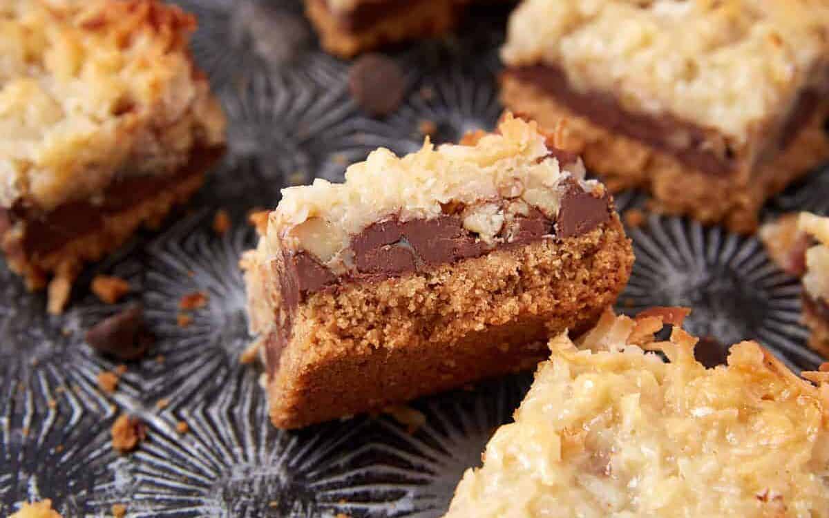 A close-up of a coconut chocolate bar with a crumbly base, a layer of chocolate, and a toasted coconut topping, set on a textured dark surface. Other similar bars are partially visible in the background.