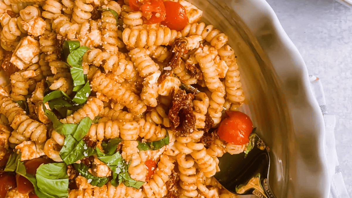 A close-up of a bowl of rotini pasta topped with tomato sauce, cherry tomatoes, sun-dried tomatoes, feta cheese, and fresh basil, with a fork resting in the dish.
