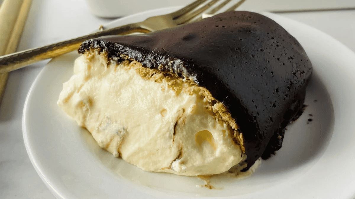 A slice of chocolate eclair cake with creamy filling and a thick chocolate topping is served on a white plate with a fork in the background.