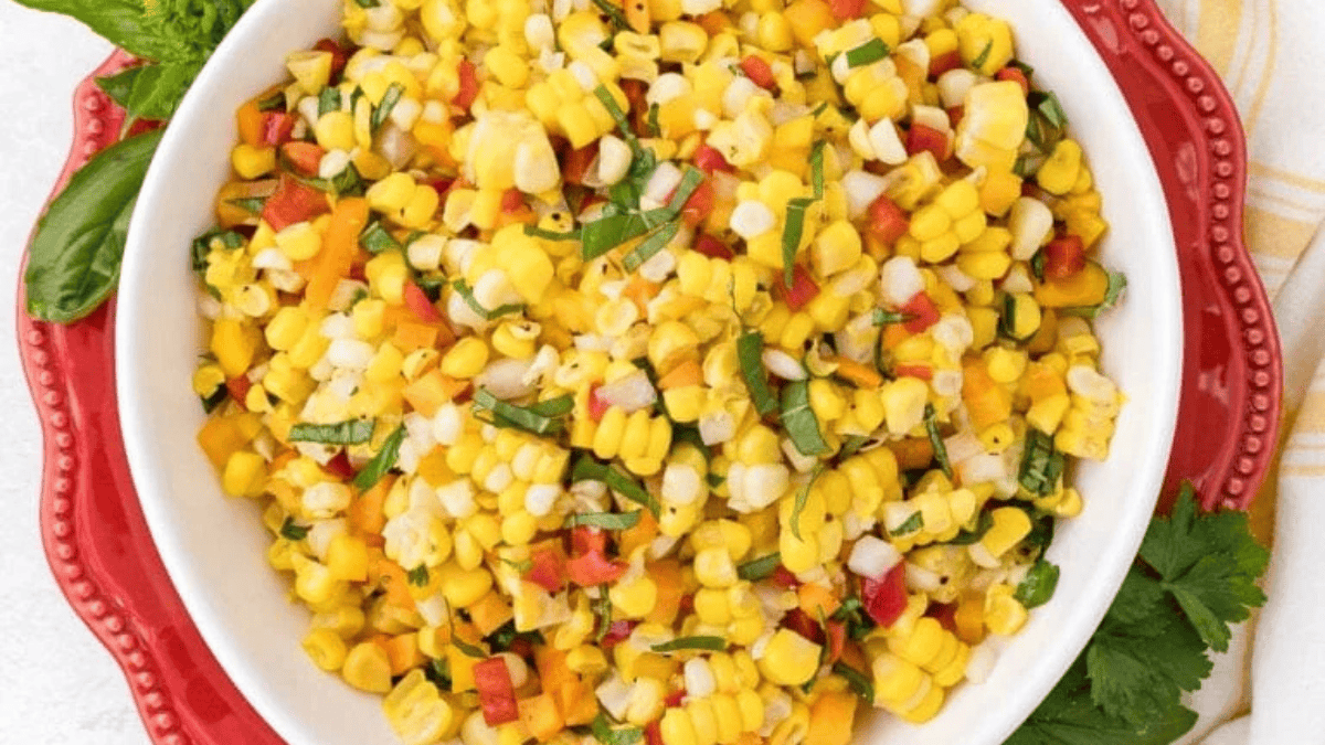 A white bowl filled with colorful corn salad, featuring yellow corn, red bell peppers, and green herbs, sits on a red plate with fresh herbs on the side.