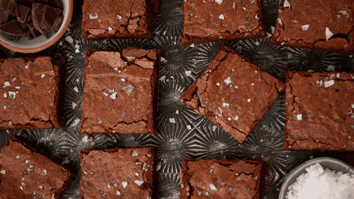 Nine chocolate brownies topped with flaky sea salt are arranged on a patterned metal surface, with small bowls of chocolate pieces and sea salt visible at the corners.