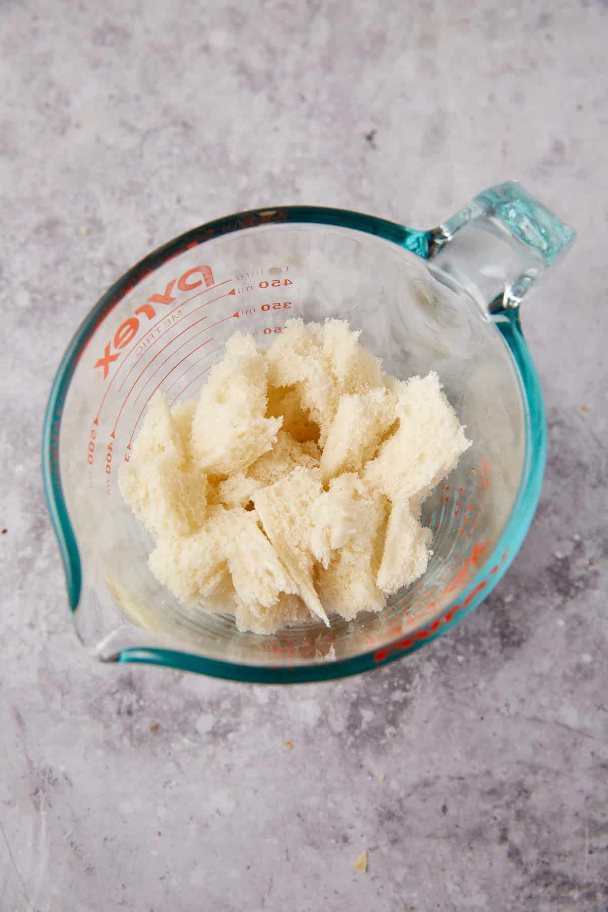 Torn pieces of white bread are placed in a glass Pyrex measuring cup on a gray, textured surface—perfect for prepping classic recipes like Grape Jelly Meatballs.