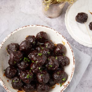 A white bowl filled with glazed Grape Jelly Meatballs garnished with chopped herbs sits on a light gray napkin. Nearby, a small white plate holds two more meatballs with cocktail sticks, and a small metal pot with sprouts is visible in the background.