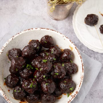 A white bowl filled with glazed Grape Jelly Meatballs garnished with chopped herbs sits on a light gray napkin. Nearby, a small white plate holds two more meatballs with cocktail sticks, and a small metal pot with sprouts is visible in the background.
