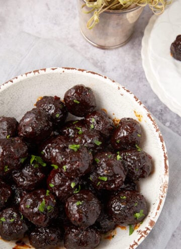 A white bowl filled with glazed Grape Jelly Meatballs garnished with chopped herbs sits on a light gray napkin. Nearby, a small white plate holds two more meatballs with cocktail sticks, and a small metal pot with sprouts is visible in the background.