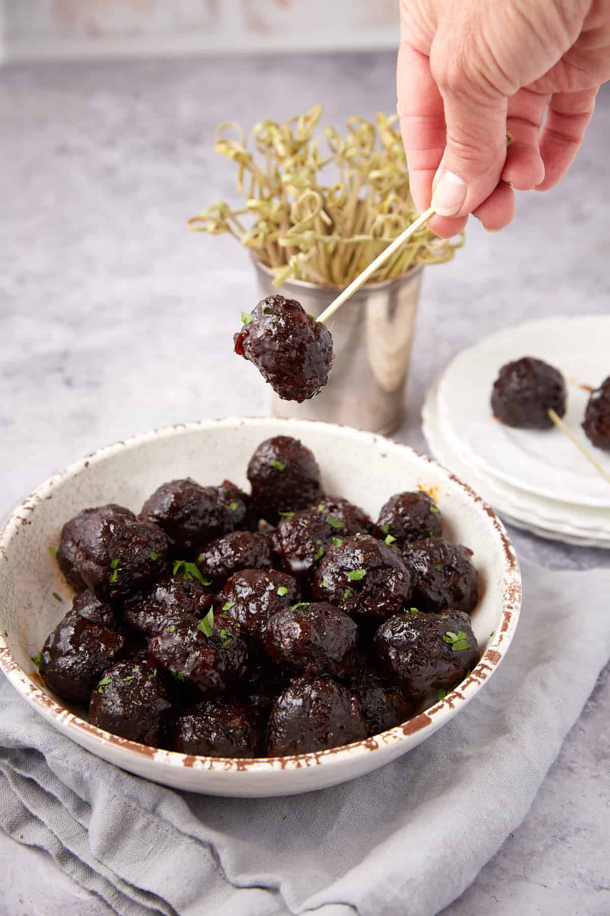 A hand holds a toothpick with a Grape Jelly Meatball above a bowl filled with more glazed meatballs, garnished with herbs. Plates and extra toothpicks are in the background on a gray surface.