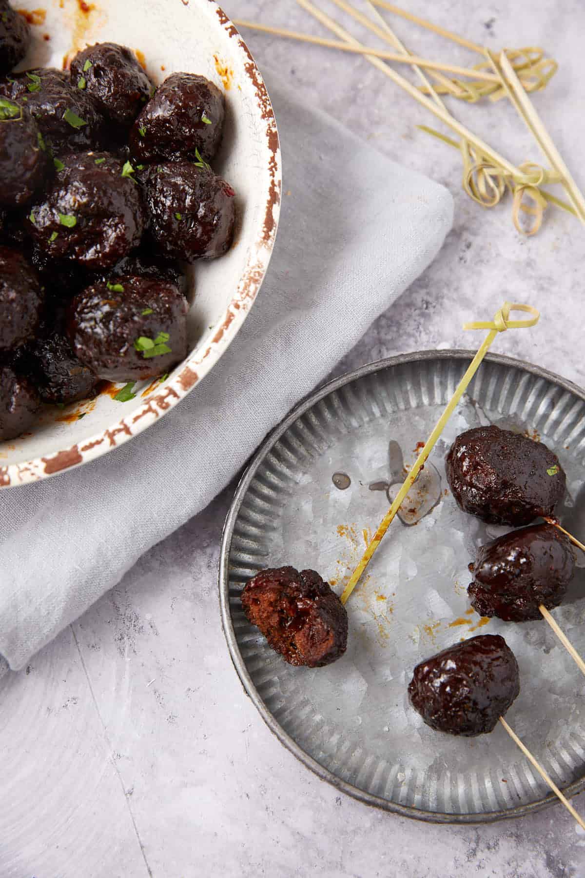 Overhead view of Grape Jelly Meatballs on wooden skewers, some on a metal plate and others in a white dish, with a light gray napkin and extra skewers arranged on a marble surface.
