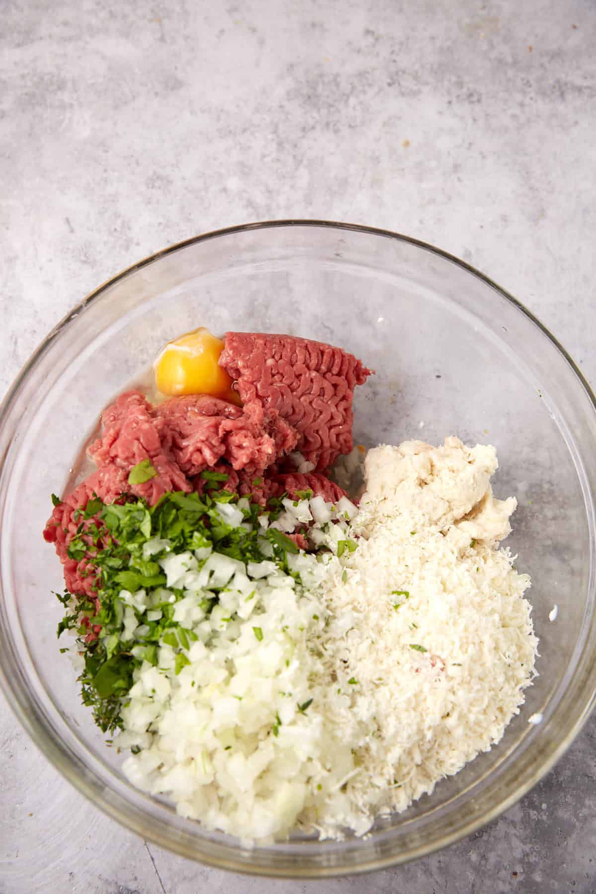A glass bowl containing ground beef, an egg yolk, chopped parsley, diced onions, breadcrumbs, and white bread on a gray countertop—perfect foundations for classic meatballs or creative dishes like Grape Jelly Meatballs.
