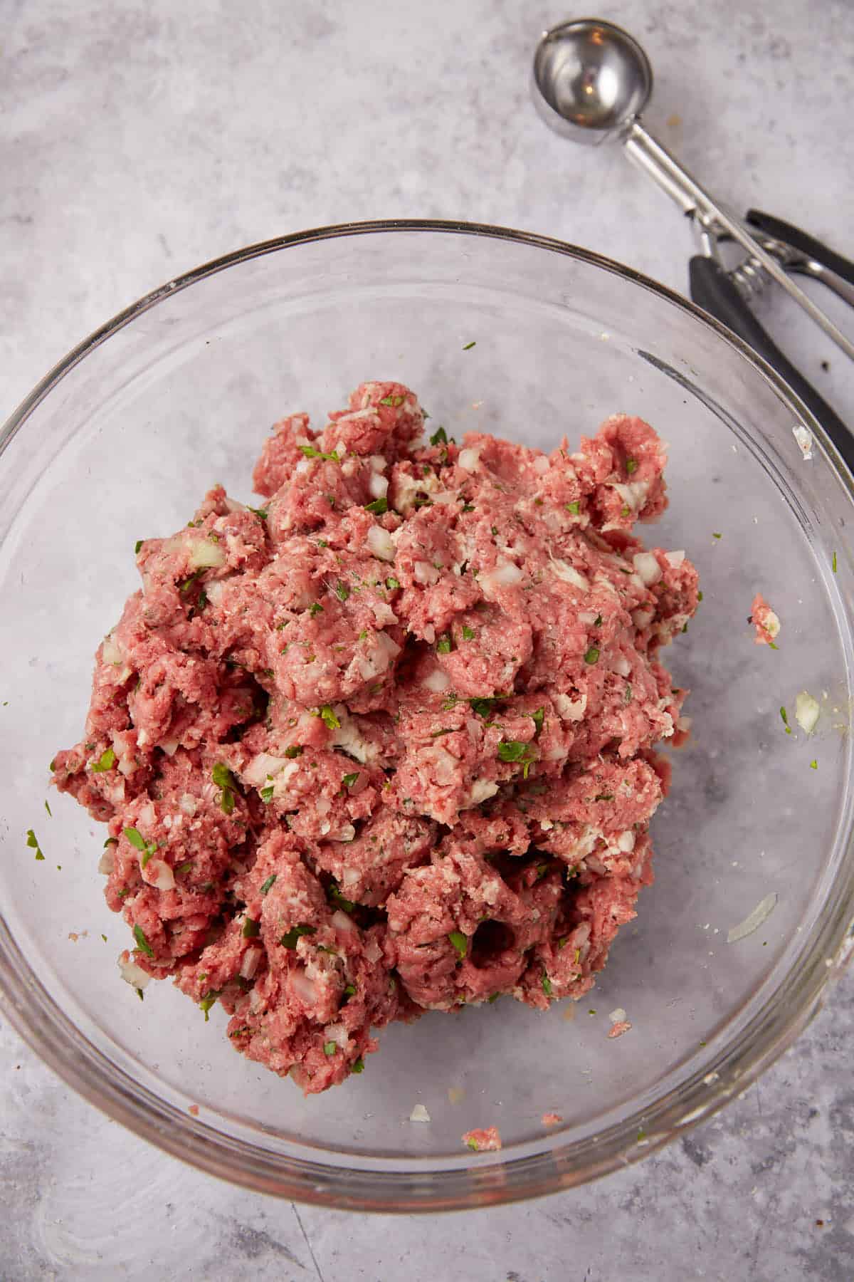 A glass bowl filled with seasoned raw ground meat mixture for Grape Jelly Meatballs, with visible chopped onions and herbs. Metal scoops rest on the gray countertop beside the bowl.