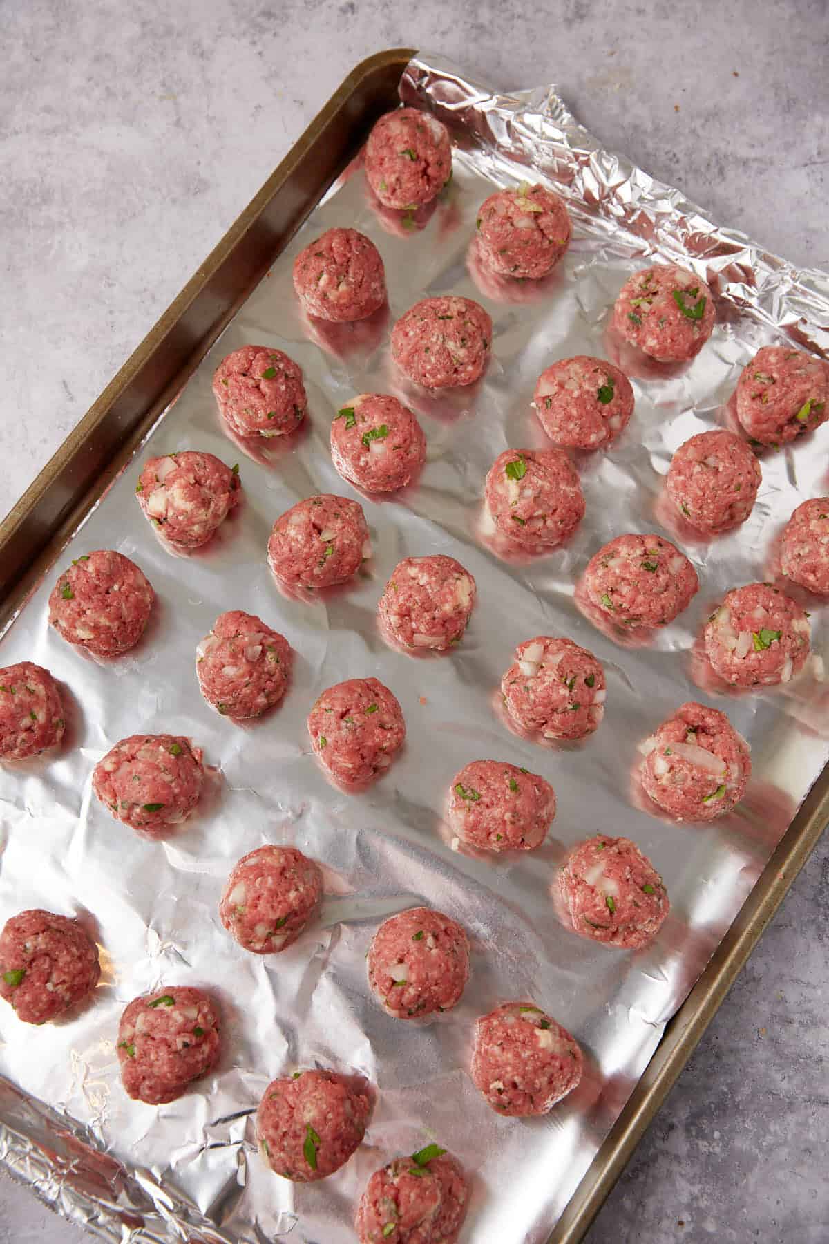 A baking tray lined with aluminum foil holds rows of raw grape jelly meatballs, mixed with herbs and onions, ready for cooking. The tray sits on a light gray countertop.
