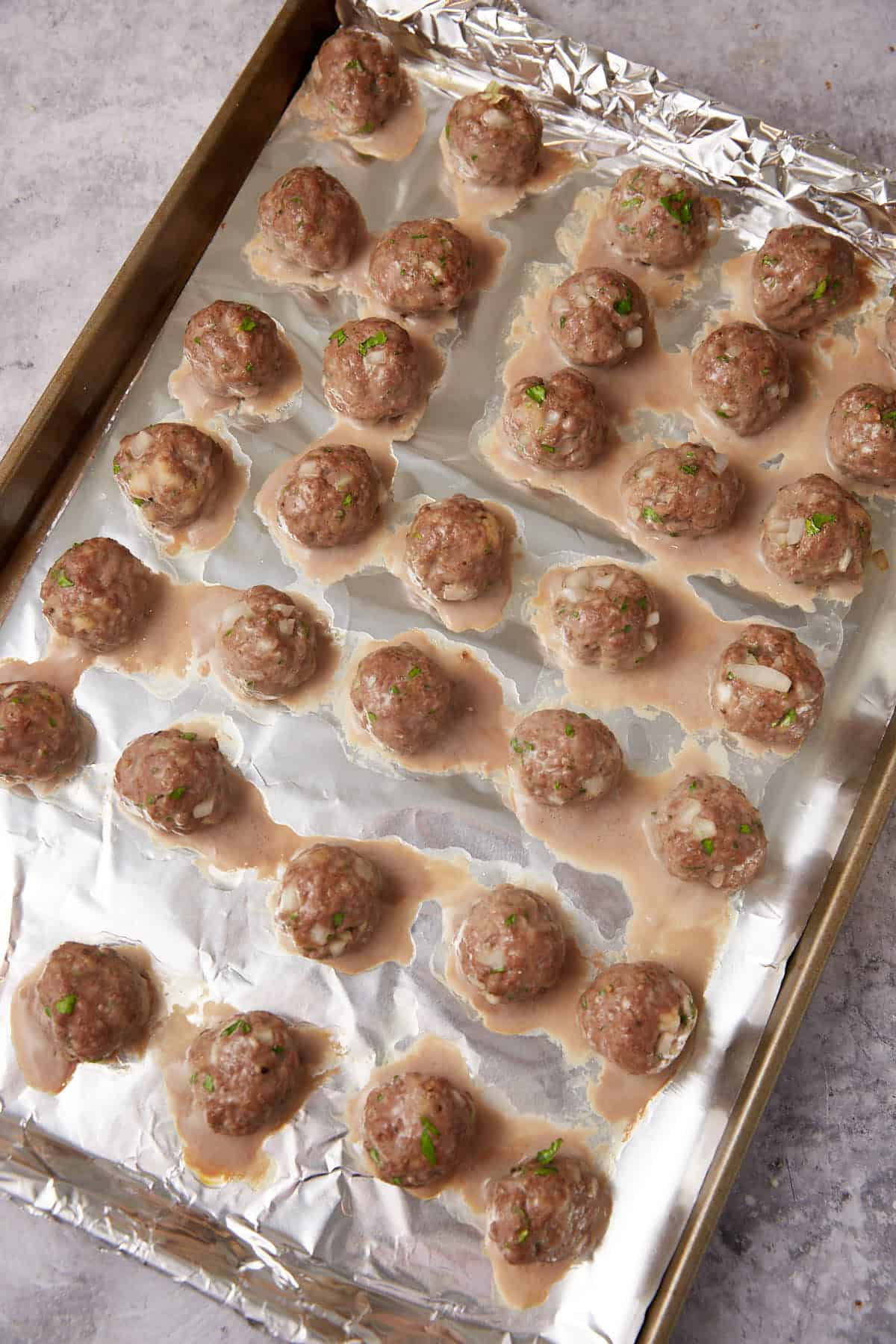 A baking sheet lined with aluminum foil holds rows of cooked grape jelly meatballs, resting in small puddles of juices. The meatballs are speckled with green herbs and bits of onion.