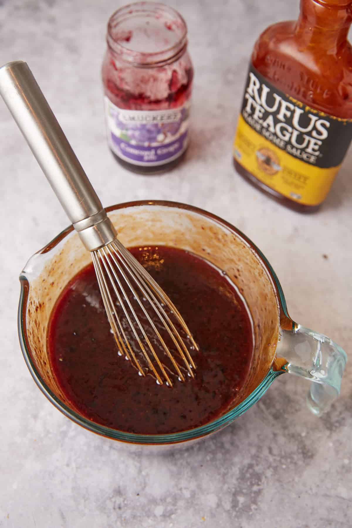 A glass measuring cup with a metal whisk inside holds a dark reddish sauce mixture, perfect for Grape Jelly Meatballs. Behind it are an open jar of blackberry preserves and a bottle of Rufus Teague barbecue sauce.