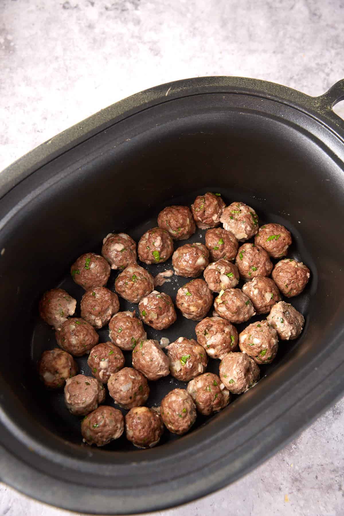 A black slow cooker filled with savory grape jelly meatballs, each cooked to perfection and garnished with chopped herbs, arranged in a single layer on the bottom of the pot.