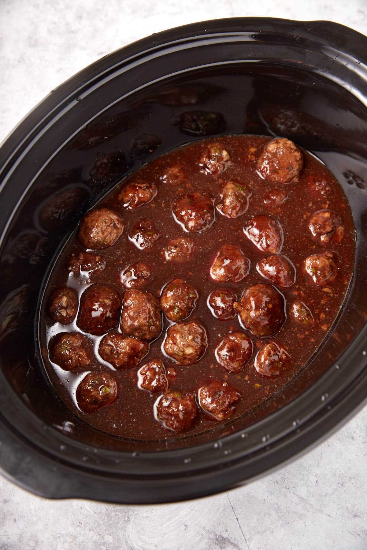 Grape Jelly Meatballs simmer in a rich, dark red sauce inside a black slow cooker, photographed from above on a light gray surface.