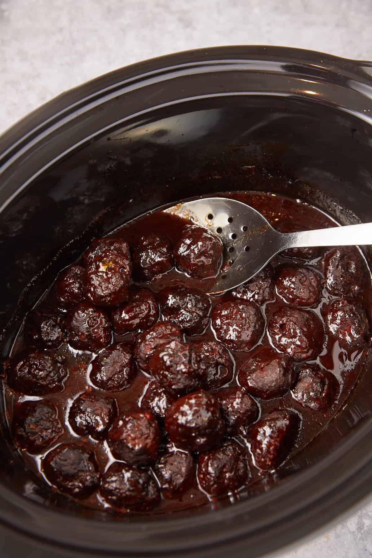 A black slow cooker filled with saucy Grape Jelly Meatballs, with a slotted metal spoon resting inside, sits on a light gray surface.
