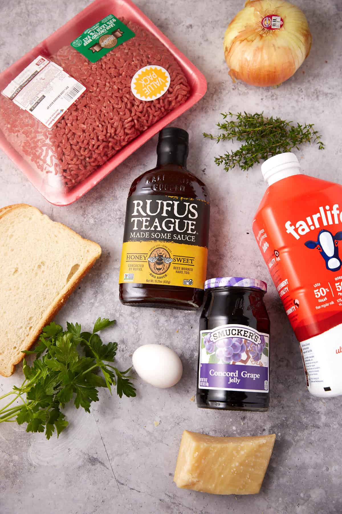 Flat lay of various food items on a gray surface, including ingredients for Grape Jelly Meatballs such as ground beef, yellow onion, barbecue sauce, whole milk, white bread, fresh herbs, a raw egg, grape jelly, and Parmesan cheese.