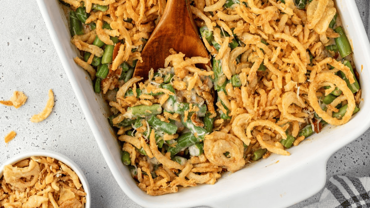 A casserole dish filled with green bean casserole topped with crispy fried onions, with a wooden spoon serving some out. A small bowl of extra fried onions is nearby on a gray surface.