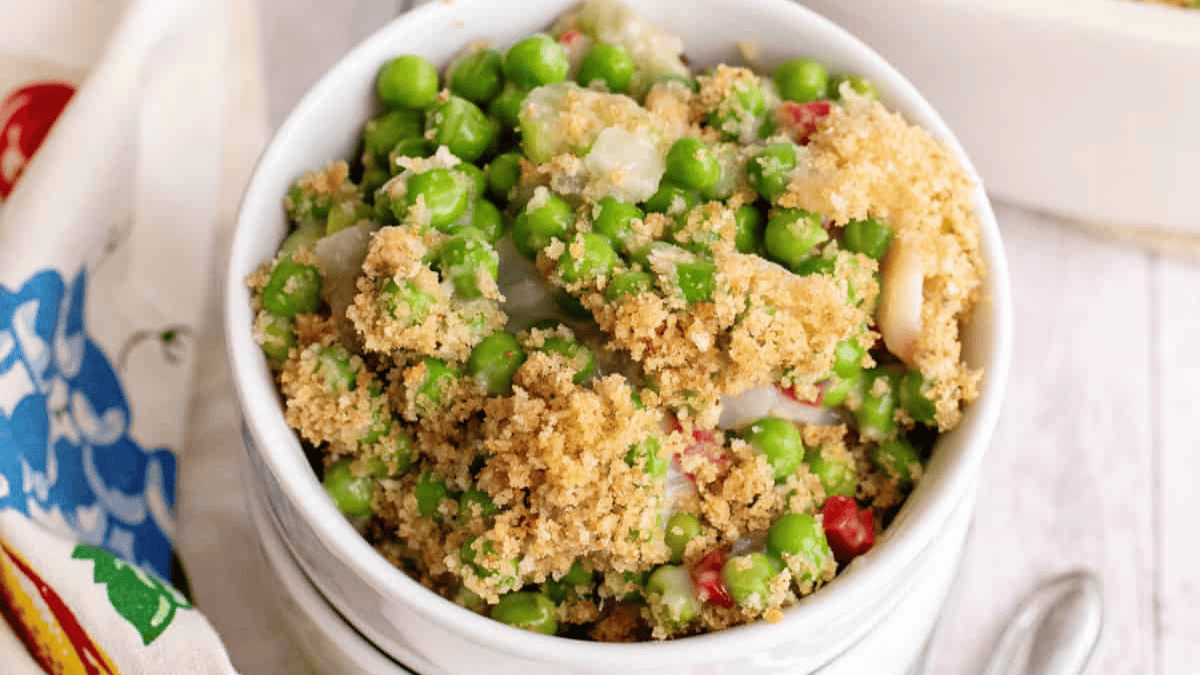 A bowl filled with a casserole made of green peas, breadcrumbs, and small diced vegetables, served on a light wooden surface with part of a patterned napkin visible on the side.