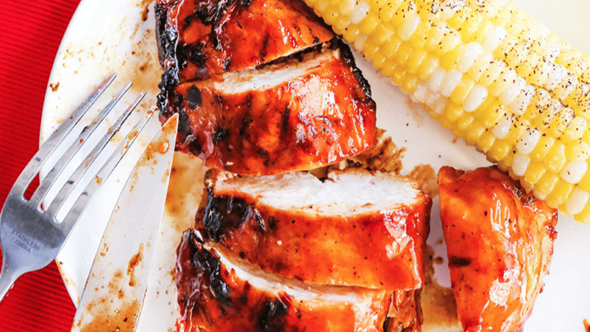 A close-up of sliced barbecued chicken breast with a glaze, a piece of corn on the cob seasoned with pepper, and a fork and knife on a white plate—perfect inspiration for your next grilling recipes.