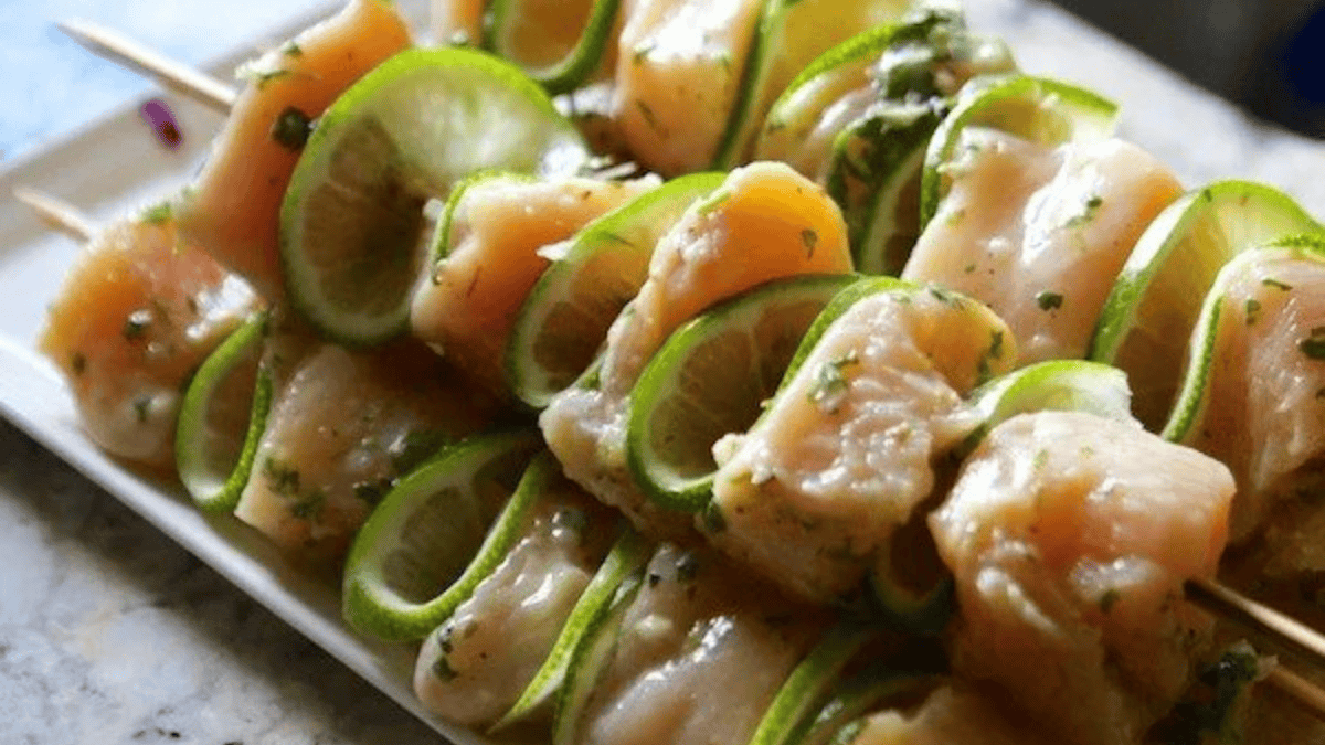 Raw chicken pieces and thin lime slices are threaded onto wooden skewers, arranged neatly on a white rectangular plate—perfect for adding to your favorite grilling recipes.
