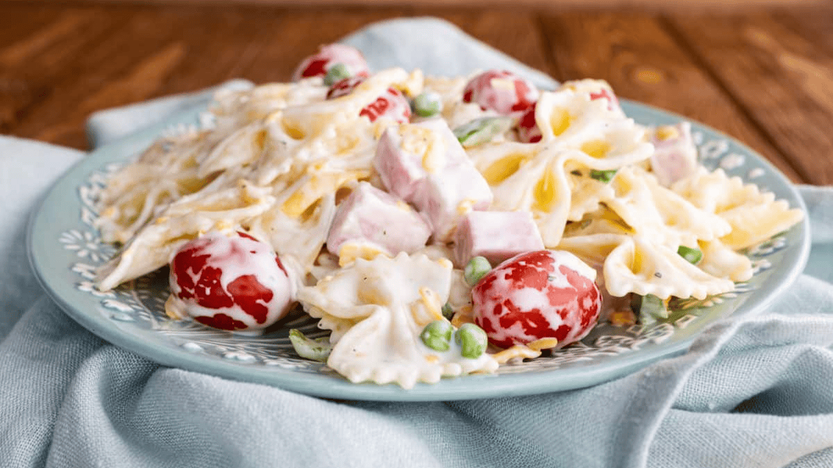 A plate of creamy pasta salad with bowtie noodles, cherry tomatoes, peas, and cubes of ham, served on a blue-green plate with a light blue cloth underneath.