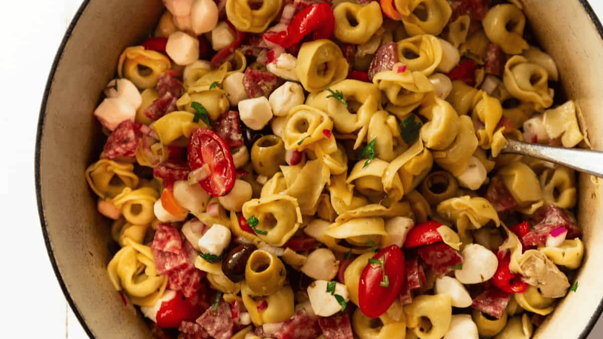A close-up of a bowl filled with tortellini pasta salad, featuring cheese tortellini, cherry tomatoes, mozzarella balls, salami slices, black olives, red onion, and fresh herbs. A spoon is partially visible on the right side.