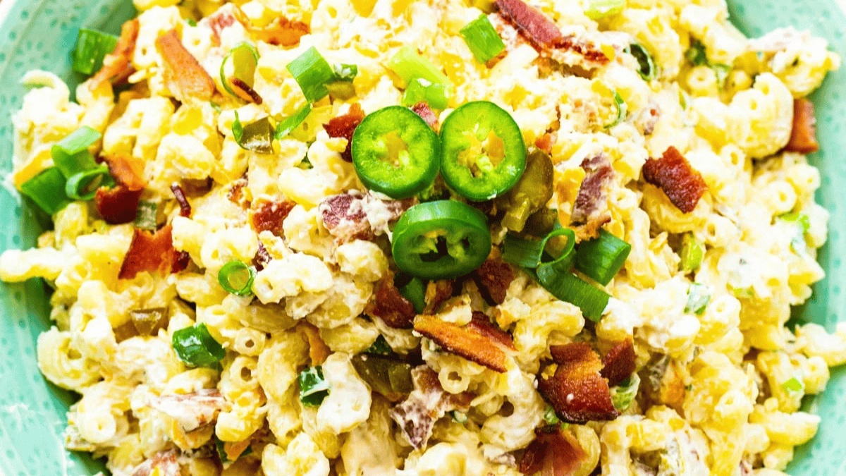 A close-up of macaroni salad topped with sliced jalapeños, crispy bacon pieces, and chopped green onions on a green plate. The salad appears creamy with a mix of pasta, vegetables, and dressing.