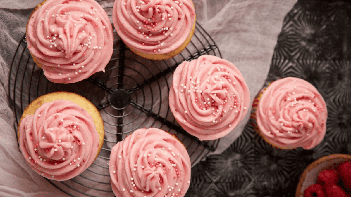 Six vanilla cupcakes with pink swirled frosting and white sprinkles are arranged on a round cooling rack, making the perfect sweet treats for mom, with part of another cupcake and a dark patterned surface visible on the side.