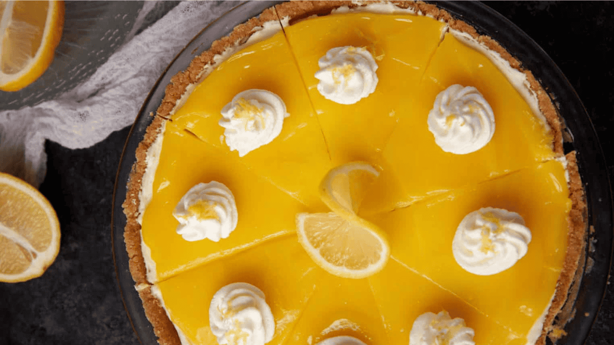 A lemon pie with a bright yellow filling, topped with whipped cream dollops and a lemon slice in the center, is sliced and displayed on a dark surface with a lemon wedge beside it.