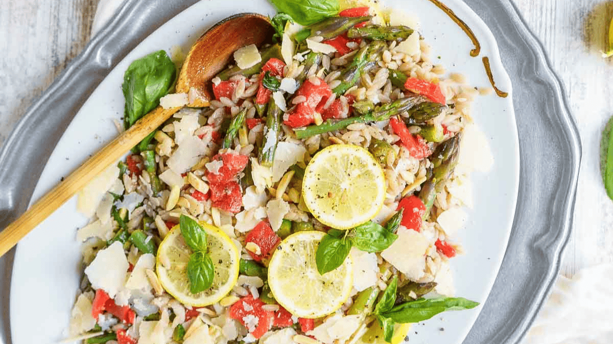 A platter of orzo salad with asparagus, diced tomatoes, shaved parmesan, fresh basil, and lemon slices, served with a wooden spoon on a silver tray.