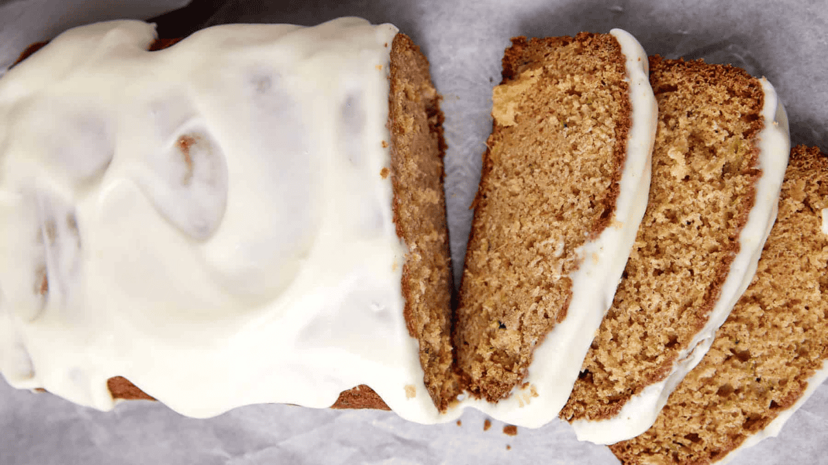 A close-up of sliced zucchini bread topped with a thick layer of white cream cheese frosting, displayed on parchment paper.