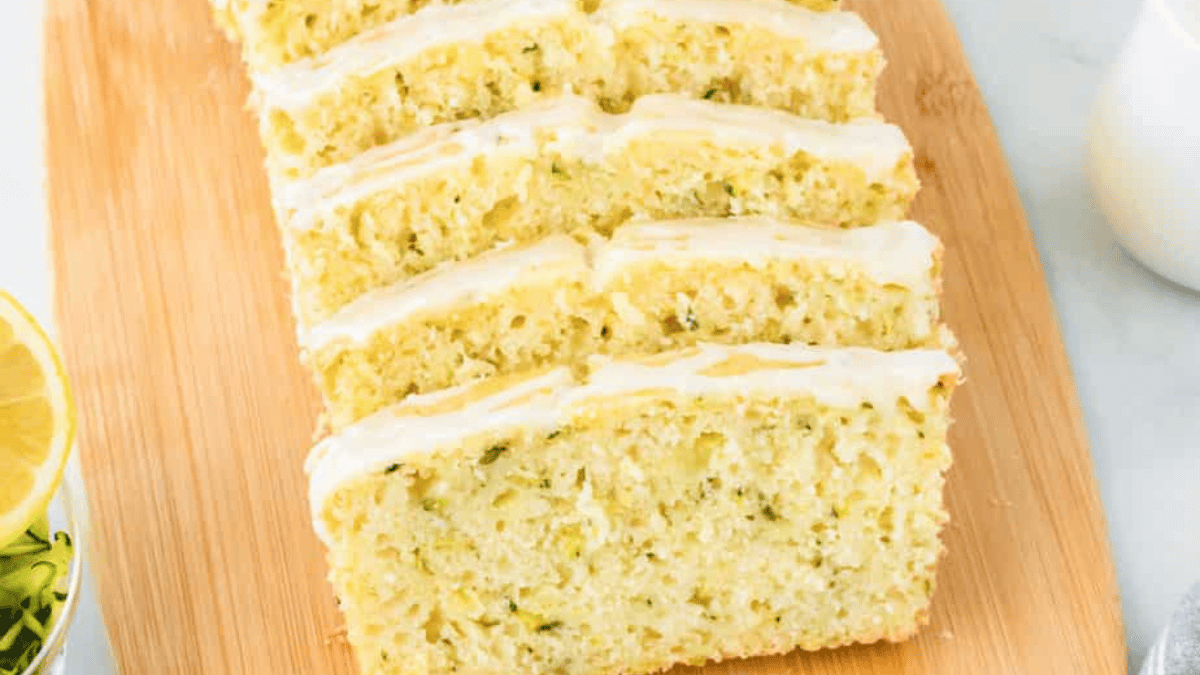 Five slices of lemon zucchini bread with white icing are neatly arranged on a wooden cutting board, with a partial lemon and a glass of milk visible in the background.