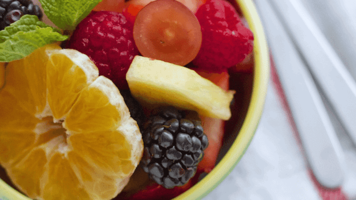 A close-up of a colorful fruit salad featuring orange slices, raspberries, blackberries, grapes, pineapple, and a sprig of mint in a bowl, with silverware visible in the background.