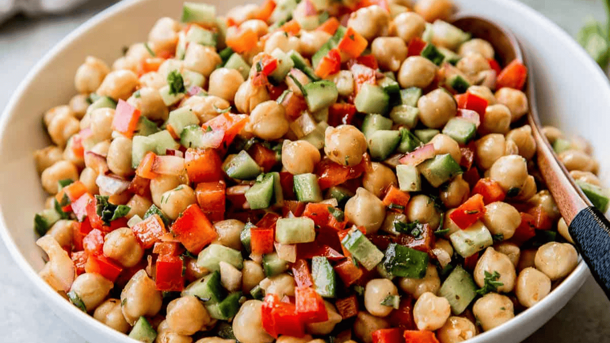 A bowl of chickpea salad with diced cucumber, red bell pepper, and herbs, mixed together and served with a wooden spoon.