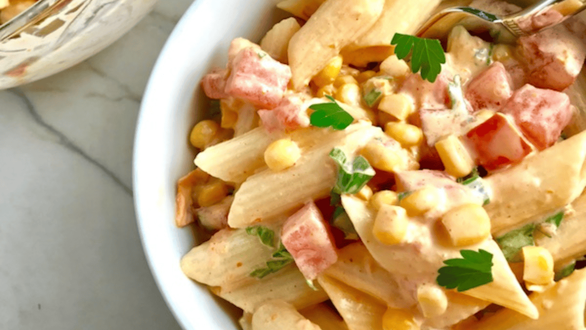 A bowl of creamy pasta salad with penne, corn, diced tomatoes, green herbs, and a creamy dressing, garnished with parsley. A fork rests in the bowl on a light-colored surface.