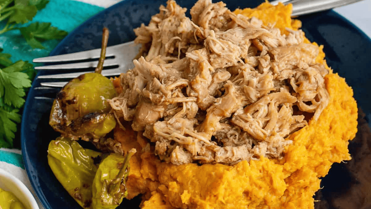 Shredded pork served on a mound of mashed sweet potatoes, with a few cooked green peppers on the side, arranged on a dark blue plate with a fork.