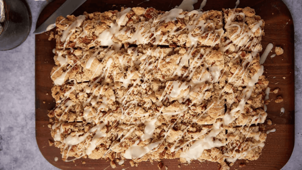 A large rectangular coffee cake topped with crumbly streusel and drizzled with white icing, sliced into squares, displayed on a wooden cutting board.