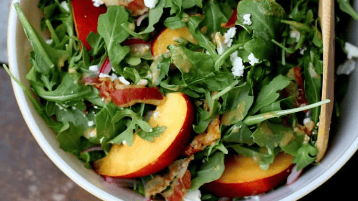 A bowl of fresh salad with arugula, peach slices, crumbled cheese, red onions, and a wooden spoon, ready to serve.