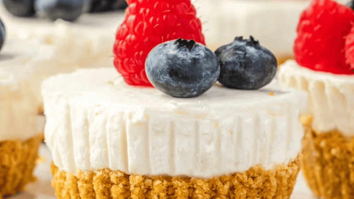 A mini cheesecake with a graham cracker crust, topped with fresh raspberries and blueberries, shown in close-up with other similar cheesecakes blurred in the background.