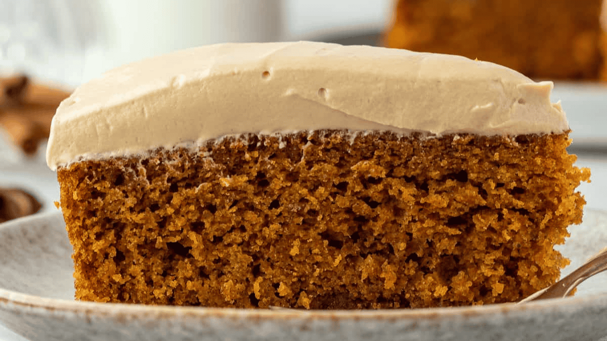A close-up of a slice of moist, spiced cake with a thick layer of creamy frosting on top, served on a light-colored plate.