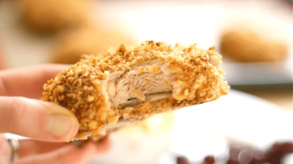A hand holds a piece of breaded and baked chicken with a creamy filling inside, showing the crispy coating and tender meat. The background is blurred with more food visible.