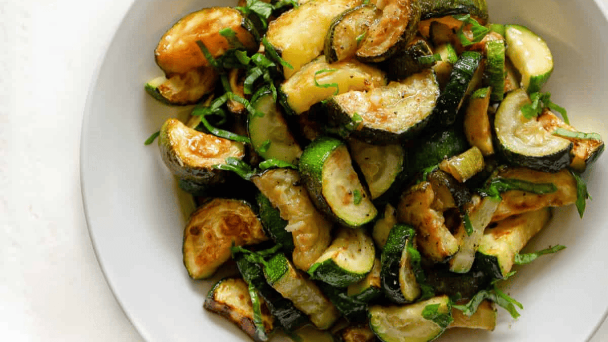 A white plate filled with sautéed zucchini slices, garnished with chopped herbs, sits on a light surface. The zucchini appears lightly browned and seasoned.