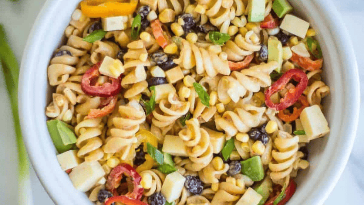 A bowl of pasta salad with rotini noodles, diced avocado, cubes of cheese, sliced mini peppers, corn, black beans, and chopped green onions.
